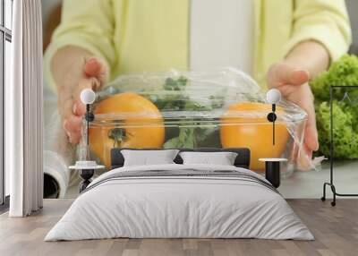 Woman putting plastic food wrap over glass container with fresh vegetables at white marble table, closeup Wall mural