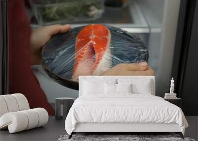 Woman putting plate of fresh salmon with plastic food wrap into refrigerator, closeup Wall mural