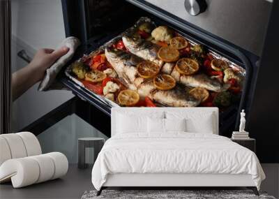 Woman taking out baking tray with sea bass fish and vegetables from oven, closeup Wall mural