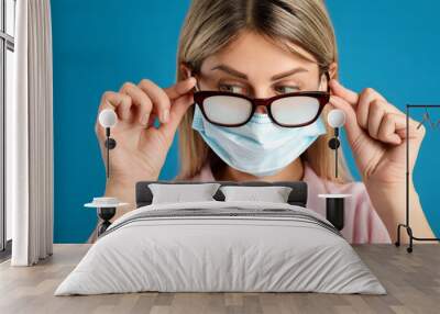 Woman with foggy glasses caused by wearing disposable mask on blue background. Protective measure during coronavirus pandemic Wall mural