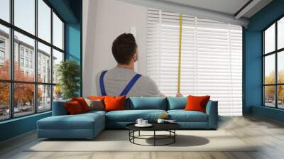 Worker in uniform using measuring tape while installing horizontal window blinds indoors Wall mural