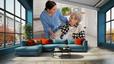 Young caregiver assisting senior woman in wheelchair indoors. Home health care service Wall mural