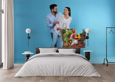 Young couple with shopping cart full of groceries on light blue background Wall mural