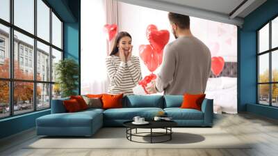 Young man presenting gift to his girlfriend in bedroom decorated with heart shaped balloons. Valentine's day celebration Wall mural