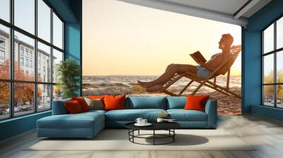 Young man reading book in deck chair on beach Wall mural