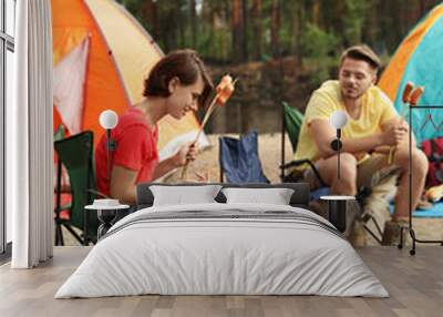 Young people having lunch with sausages near camping tents outdoors Wall mural