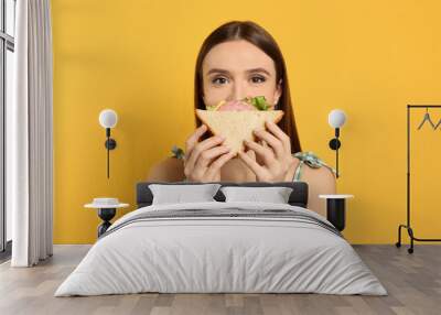 Young woman eating tasty sandwich on yellow background Wall mural