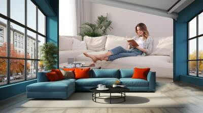 Young woman reading book on sofa at home Wall mural