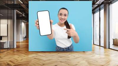 Young woman showing smartphone in hand and pointing at it on light blue background Wall mural