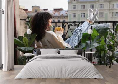 Young woman with cup of tea relaxing in chair surrounded by green houseplants on balcony Wall mural