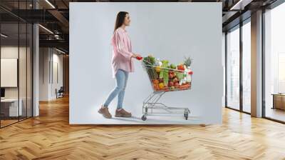 Young woman with shopping cart full of groceries on grey background Wall mural