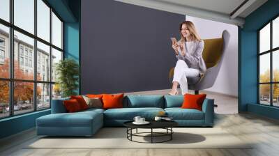 Young woman with smartphone sitting in armchair at home. Space for text Wall mural