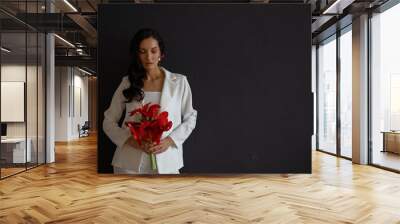 Fashion portrait of young woman poising in white trouser suit. Wedding trends and concept. Holding bouquet of red amaryllis. Dark background Wall mural