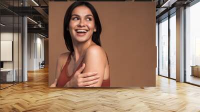 Laughing vivacious young woman with a beaming smile embracing herself with her hands while posing over brown background. Happy woman with naked shoulders laughing and looking away Wall mural