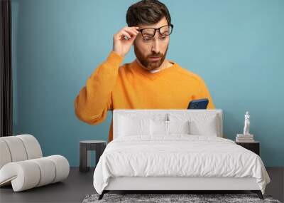 Mobile connection. Portrait of shocked astonished man reading post on social network using cell phone, chatting looking surprised. Indoor studio shot isolated on blue background Wall mural