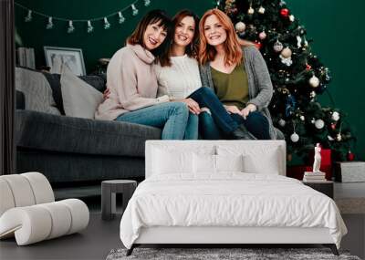 Best friends forever. Portrait of three attractive middle aged women seated together on a sofa with a Christmas tree in the background at home. Wall mural