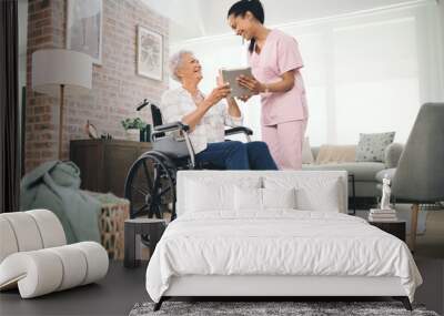 Ive captured all your big moments. Shot of a young nurse sharing information from her digital tablet with an older woman in a wheelchair. Wall mural