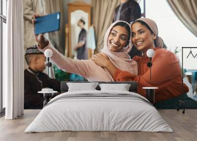 Muslim sisters taking selfies in traditional Islamic head scarf inside a happy family home together. Beautiful young women who are proud of their religion post photos of Eid Ramadan on social media Wall mural