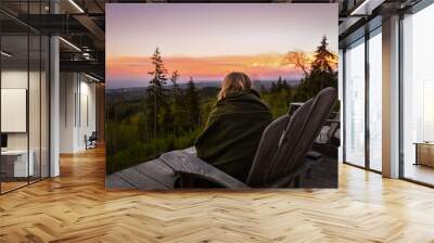 Woman in blanket on wooden chair watches sun rise over forest Wall mural