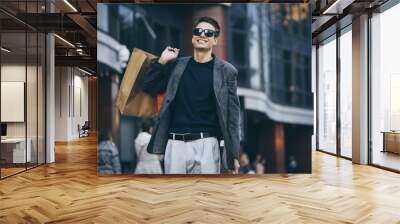 Serious stylish young man with sunglasses walking in urban street and enjoying Black Friday shopping in trendy stores in city Wall mural