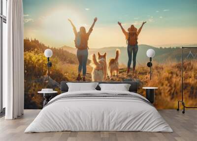 Two hikers hiking on mountain pathway with two dogs during sunset female friends hands in the air happy celebrate success reach destination Wall mural