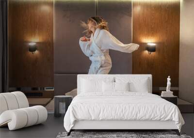 Woman having fun and jumping with bathrobe on bed in hotel room Wall mural