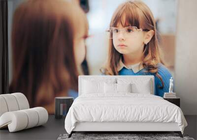 Little Girl Wearing New Eyeglasses Looking in the Mirror. Adorable fashionable toddler wearing stylish frames in optical store
 Wall mural