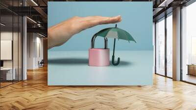 Hand holds pink padlock under green umbrella on light blue surface, symbolizing security Wall mural