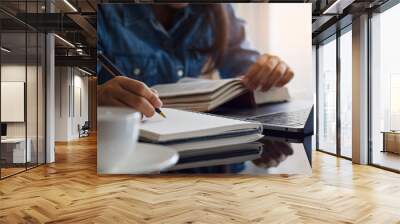 Casual young female student handwriting information on diary notebook while reading book and work on laptop computer with white cup of coffee on the table at home. Wall mural