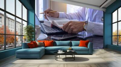 Male doctor in white lab coat with stethoscope concentrated on reading book, work on laptop computer and mobile smart phone on the desk.	 Wall mural