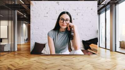 Authentic lifestyle portrait of attractive young caucasian woman in eyeglasses sitting on a sofa at home and looking at camera. Wall mural