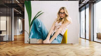 Portrait of attractive caucasian young woman in eyeglasses sitting on chair near white wall. Wall mural