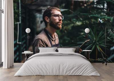 Young man gardener in glasses and apron with digital tablet working in a garden center for better quality control. Environmentalist using digital tablet in greenhouse. Wall mural