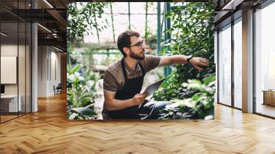 Young man gardener in glasses and apron with digital tablet working in a garden center for better quality control. Environmentalist using digital tablet in greenhouse. Wall mural