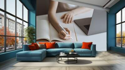 Young woman works on a laptop and takes notes in a notebook. Work in home office. Wall mural