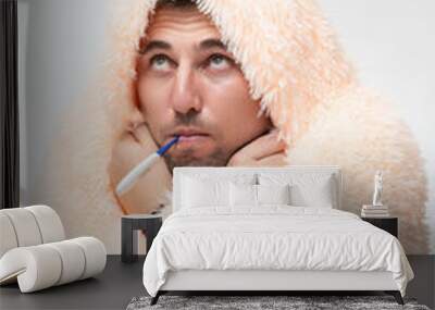 A sick man wrapped in a blanket holds a thermometer in his mouth. Wall mural