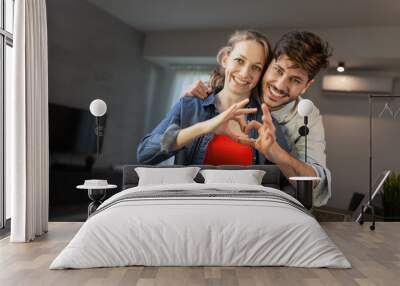 A joyful young couple forms a heart shape with their hands, smiling warmly in their modern living room with a laptop in the background. Wall mural