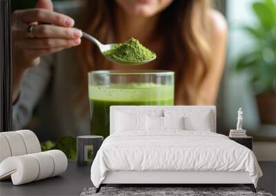 A woman carefully adds vibrant green matcha powder into a glass of blended smoothie, highlighting the preparation of a healthy, energizing drink. Wall mural