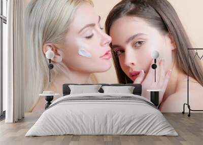 Skin care together. Girls friends relaxing with facial masks on over beige background. Applying face cream together. Blonde and brunette on beige background. Beauty procedure. Wall mural