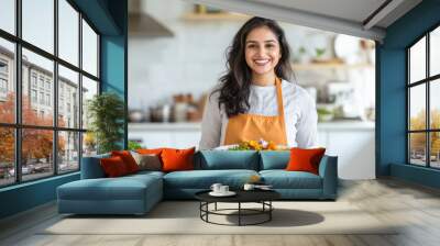 a beautiful smiling young indian woman in an apron holding a plate of food standing in a kitchen at home. Wall mural