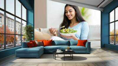 attractive indian woman standing and eating salad at the kitchen counter Wall mural