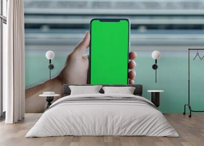 A hand holding a smartphone with a green screen, perfect for adding your own content or branding. The blurred background creates a sense of depth and space. Wall mural