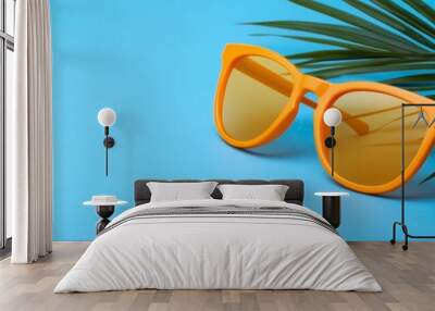 A pair of vibrant orange sunglasses rest on a bright blue background, with a palm leaf peeking into the frame. The image evokes a sense of summer, relaxation, and carefree fun, perfect for a beach vac Wall mural