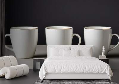 Three elegant white mugs with gold rims are lined up on a gray surface against a dark backdrop, showcasing minimalist and sophisticated design. Wall mural