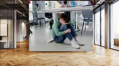 Depressed lonely boy sitting under desk in classroom with classmates playing on background Wall mural