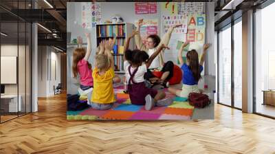 Elementary school teacher sit in class on floor with little pupils and moving hands up Wall mural