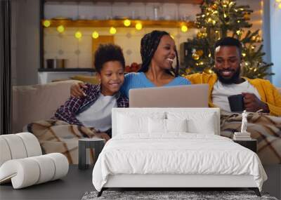 Happy African-American family resting on couch drinking tea and watching movie on laptop on christmas eve Wall mural