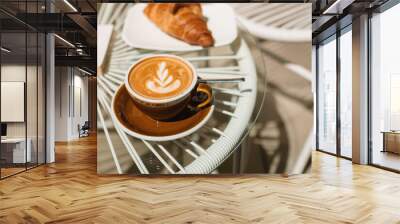 A cup of fragrant cappuccino and a warm croissant on a glass table on the terrace of the cafe. Wall mural