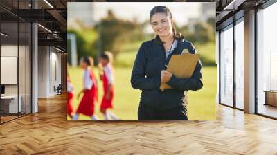 Soccer field, woman coach with and girl team training on grass in background. Sports, youth development and teamwork, a happy young female volunteer coaching football team with clipboard from Brazil. Wall mural