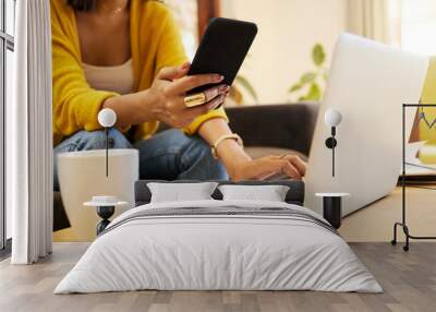 Closeup of woman working remote while typing on her laptop and holding her smartphone sitting on a sofa in a bright living room. One focused hispanic young female at home using modern technology Wall mural